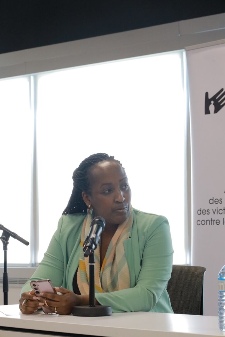 A woman in green jacket sitting at table with microphone.