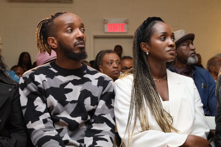 Two people sitting in a room with one of them looking at the camera.